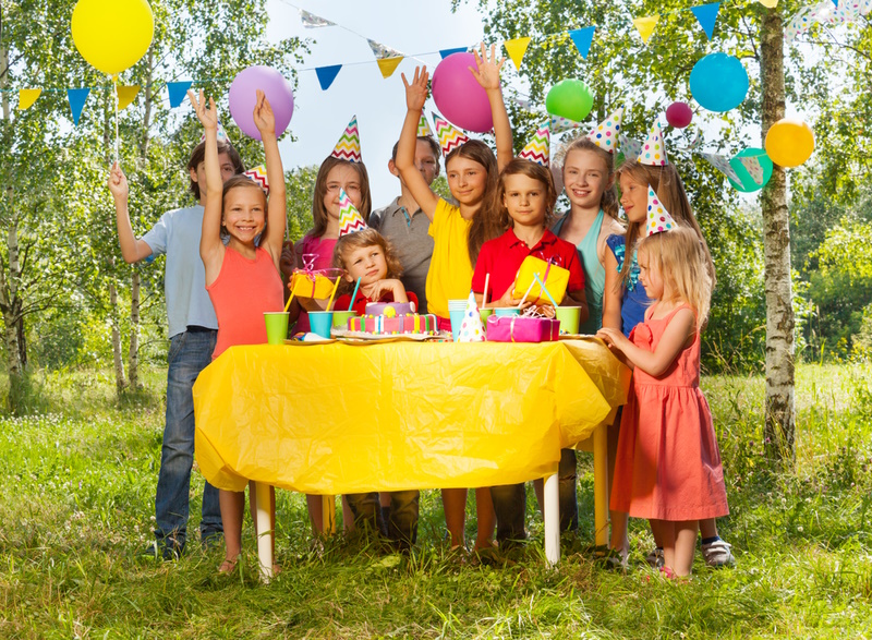 Group of happy age-diverse kids standing next to the table with Birthday cake at the outdoor B-day party